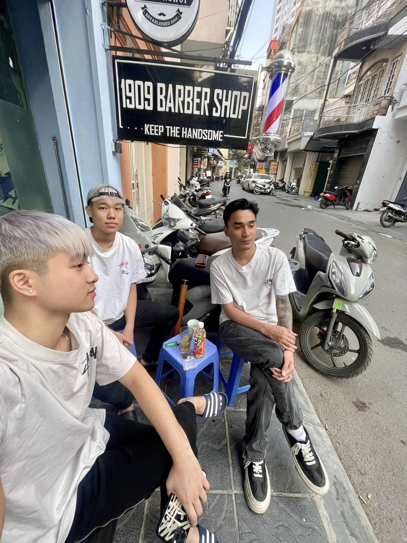 Nhóm bạn trẻ ngồi trước cửa hàng 1909 Barbershop, phía trên là bảng hiệu và cột biển báo cắt tóc